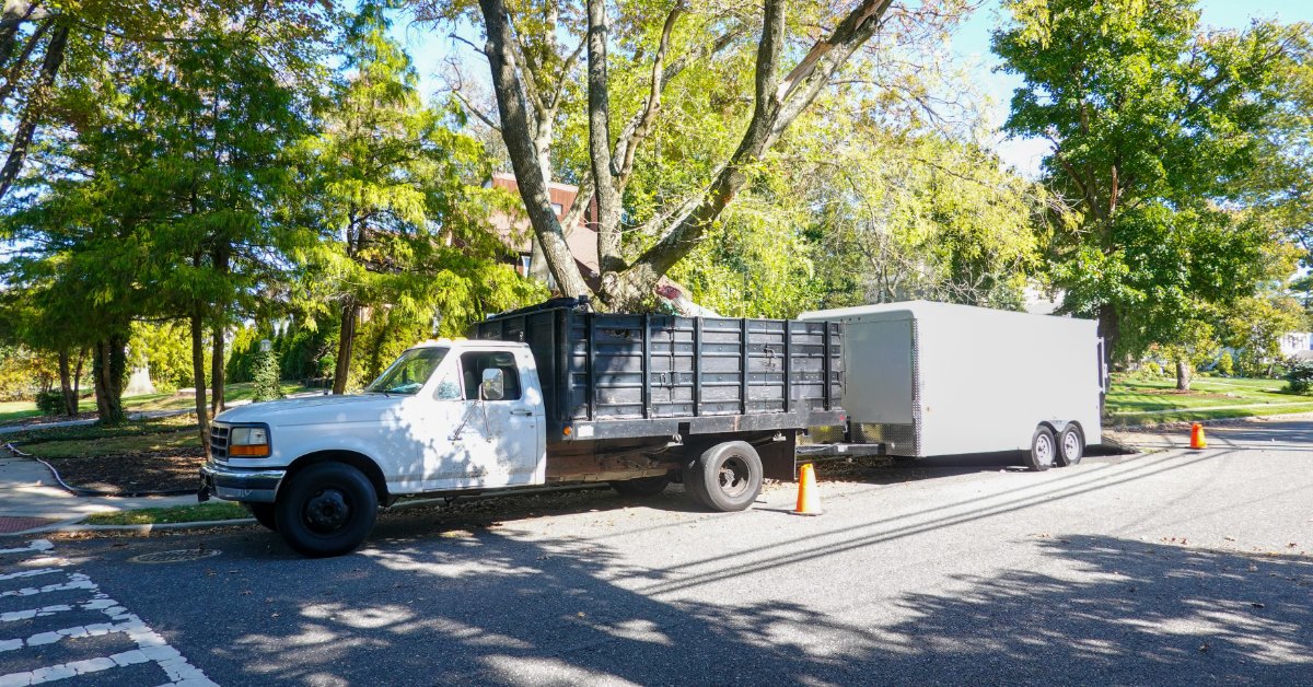 A work truck towing a white enclosed cargo trailer down a tree-lined street